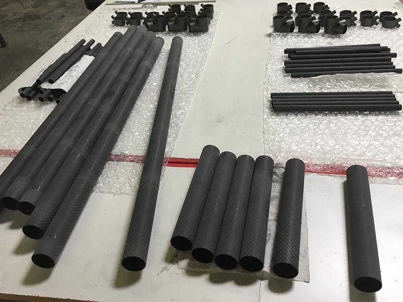 Image shows 3D printed gussets in a dark grey color and multiple carbon fiber tubes lined up in sections on a white table with plastic bubble wrap protecting them from the table surface. Materials are lined up in preparation for bonding the custom truss structure.